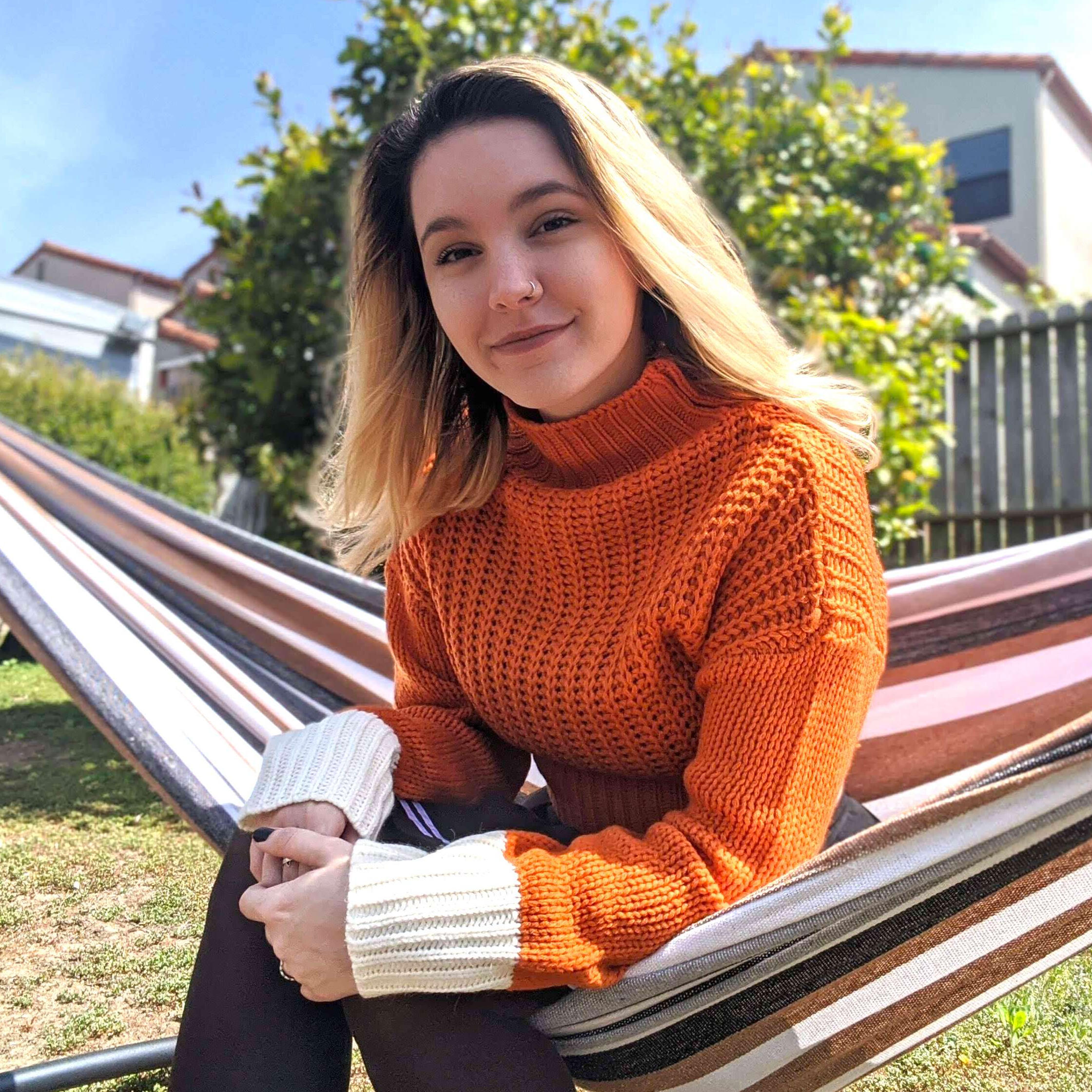 A photo of Faye Ferre smiling warmly at the camera, wearing a cozy knit orange sweater while sitting on a hammock during Golden Hour.